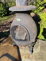 Front angle showing chimenea body with mesh door and cast iron legs, rust and weathering visible.