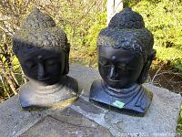Two black concrete Buddha bust statues on outdoor stone slab with moss growth