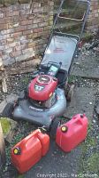 Craftsman gas lawn mower with visible engine, wheels, and safety handle, alongside three red gas cans on outdoor ground with brick wall background