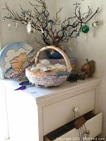 Photo showing Easter decor with woven basket of eggs and branch tree with hanging ornaments, Winnie the Pooh painted round plate on the dresser.