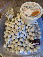 Large transparent plastic bin containing over 100 used white golf balls with some logos and markings visible. A large white ice cream bucket is also present on top within the bin.