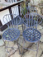 Four metal patio bar stools with lattice seats and rounded backrests, showing wear and outdoor weathering, placed on stone patio.