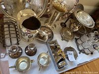 Photo showing large embossed platters, a silver plated pitcher, silver plated handled cups, candle holders, and crystal cruets in silver holder on a white surface.