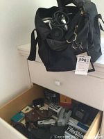 Wide view of opened drawer containing vintage cameras and accessories with a black camera bag above with cables and lenses inside, plus tag indicating lot number.