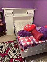 Twin bed setup with white wooden headboard attached showing storage on top and sides, twin mattress included, multiple decorative pillows and comforter arranged on the bed, purple walls and floral area rug visible.