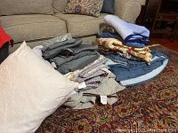 Photo of folded pillows, blankets, and bed linens stacked on a carpet with a sofa in the background