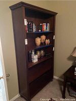 Front view of the wooden bookcase against the wall, showing three upper shelves with books and decorative vases, bottom shelf empty. Damage is visible on the lower front panel.