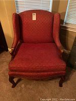 Front view of red upholstered wingback chair with wooden legs and arm accents, showing full seat cushion, fabric pattern, and nailhead trim details.