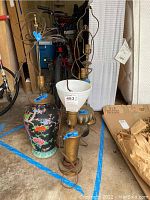 Four assorted lamps grouped on floor including large decorative ceramic lamp, two brass lamps, and small ceramic lamp with damaged cord and white shade