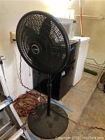 Front view of a black Lasko 20 inch diameter standing fan on a round weighted base showing the fan grille and orange extension cord coiled beside the base.