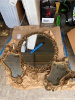 Large oval mirror lying on cardboard with detached golden wooden frame featuring floral and scrollwork designs.