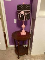 View of the pink lamp with black polka dot lampshade and wooden side table beneath it against a purple wall.
