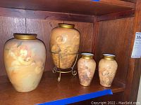 Lot of four decorated glass vases of varying sizes arranged on a wooden shelf, with one vase on a metal stand. The vases are painted with cherub or angel figures in pastel colors with gold rims.