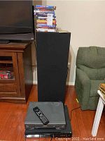 Stack of DVDs and VHS movies on top of a black speaker, with TV and furniture nearby.
