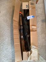 Top view showing a cardboard box containing multiple airsoft guns with visible orange muzzle tips and mix of wood and black polymer parts.