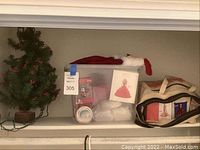 Photo of small artificial Christmas tree with red berries and attached string lights on wooden base, next to clear plastic tub with assorted Christmas decorations and canvas bag.