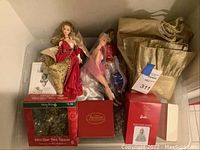 A plastic storage tub containing various Christmas decorations and two Barbie dolls, a gold fabric tree skirt, a mini star tree topper in box, and a box of outdoor light string clips.