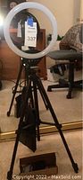 Overall view of circular LED ring light mounted on adjustable tripod stand in indoor setting with carpet and office chair background.