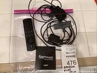 Amazon Fire TV Stick with voice remote, cables, and instruction manual arranged on a tiled surface, with the lot tag visible.