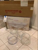 Photo of boxed ceramic twin food warmer in original packaging with a cardboard box behind it, and four clear glass bowls placed on a tiled floor in front of the box.