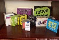 Photo showing nine boxed party/trivia games arranged on a wooden surface including Scattergories, Rotten Apples, Outburst, Things They Don't Teach You in School, What Do You Meme?, I Should Have Known That, Who in Room?, Game of Phones, and an index card holder accessory.