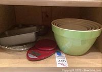 Photo of kitchen cabinet shelf with five nested green ceramic mixing bowls, red digital food scale, rectangular metal loaf pan, heart-shaped metal pan, and clear glass pie plate.