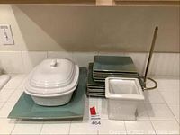 Green square dinner plates, dessert plates stacked with white ceramic covered tureen, square plant pot and gold-tone paper towel holder grouped together on countertop.