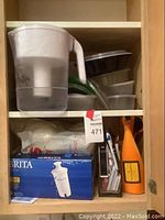 Kitchen cabinet shelf with Brita pitcher, food containers, Brita filter box, orange wine carrier and pens.
