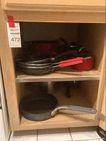 Photo showing shelf with multiple frying pans, a small red saucepan with lid, and a large frying pan with a gray handle.