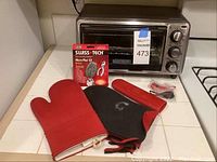 Photo showing the Black+Decker toaster oven on countertop with oven mitts, pan mitt, Swiss Tech key ring, and red bike lights placed in front as accessories.