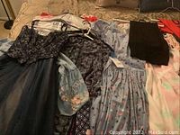 Photo showing several vintage and modern ladies dresses on hangers including a dark navy floral bodice dress with tulle skirt, blue dress with star and flag print, paisley printed dress with blue puff sleeves, and other dresses folded or hanging.