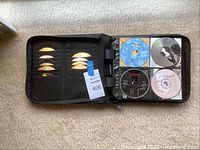 Black zippered CD organizer case opened to display multiple CDs. Left side shows ten CDs in sleeves, right side shows four CDs in clear plastic pockets with visible printed labels.