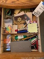 Open drawer showing mixed office supplies and stationery items including pens, pencils, glue sticks, key rings, and note pads.