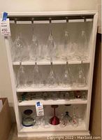 Frontal view of white wooden shelving holding 16 hanging clear wine glasses and various glassware items on bottom shelf.