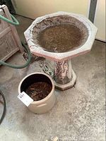 View of the cement bird bath with small cracks and the stoneware crock beside it containing leaves and debris on the floor.
