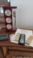 Full view of standing clock with three analog dials and Galileo thermometer alongside digital weather station, sensors and manuals.