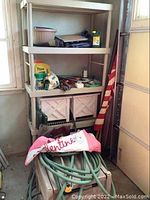 Wide view of plastic shelving unit containing hand tools, gardening supplies, and storage boxes. On the floor in front, a plastic hose caddy with green garden hose curled on top, and a folded pink flag with 'Valentine' text visible.