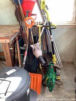 Photo showing various yard tools including Ryobi battery trimmer, pole saw leaning against wall with shovels, rakes, blower, and trash cans in front.