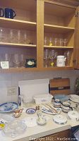 Open kitchen cabinet containing glassware including wine glasses, stemware on shelves and porcelain dishes and plates on counter with chopsticks, cutting boards, and other kitchen items.
