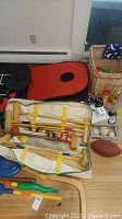 Photo showing croquet set in beige LL Bean bag, bocce balls, bean bag toss boards, brown football, bats, dice, white balls, and a wicker basket with accessories placed on floor.