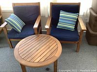 Two wooden patio chairs with navy blue cushions and striped accent pillows, plus one round wooden table with slatted top, arranged on carpeted floor near window.