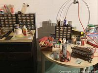Photo of black and clear plastic small-parts organizers filled with screws, nails and other hardware items, joint compound containers, hand tools, and tapes on two tables in a basement near a wall with pipes.