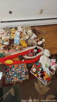 Overview of collection of various stuffed animals sitting on a wooden floor against baseboard heater, and box of small Smurf figurines on carpet