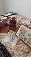 Overview photo showing five needlework pieces including woven textile with southwestern figures, floral embroidery, religious cross stitch, Eiffel Tower embroidery, all displayed on a patterned couch.