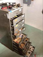 Metal cabinet with drawers open, containing assorted small tools and hardware, and various items arranged on top including power tool, level, and magnifying light.