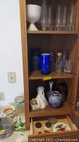 Photo of glassware cabinet including the blue blown vase, corked bottles, clear glass vases, and pottery figurines including a lady planter and other ceramics.