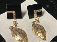 Black enamel square studs laid beside leaf‐shaped gold dangles on white paper