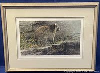 Framed print showing the full artwork with raccoon on rocky landscape, framed with beige mat and wooden frame.