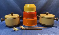 Two beige and brown enamel covered pots with lids and wooden side handles, three nesting mixing bowls stacked inside each other in orange and yellow colors, and a pair of rost-frei salad tongs with wooden handles placed in front.