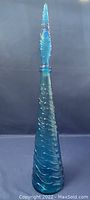 Tall blue glass genie bottle decanter with raised wave pattern, standing upright with pointed stopper in place.